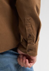 Bruine, gestructureerde shirt met een knoopmanchet en driehoekige stikdetails. Het materiaal lijkt zacht en warm. Een hand die zichtbaar een denim zak vasthoudt.