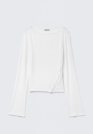 Chemise blanche à manches longues avec un col bateau et des boutons-pression diagonaux sur le bas côté droit, étiquette de la marque « Weekday » à l'intérieur du col.