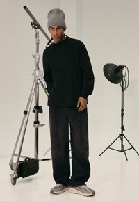 Black long-sleeve shirt and dark pants with distressed edges, paired with gray sneakers. A gray beanie completes the outfit. Studio backdrop.