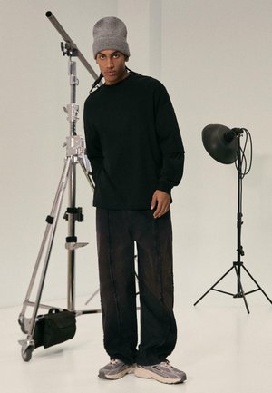 Fekete, hosszú ujjú ing és sötét nadrág, koptatott szélekkel, szürke sportcipővel párosítva. Egy szürke sapka teszi teljessé a szettet. Stúdió háttér.