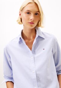 Blonde Frau mit goldenen Creolen und einem blauen und weißen gestreiften Hemd mit kleiner Logo auf weißem Hintergrund.