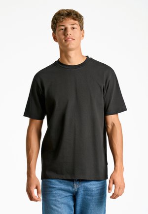 Zwarte T-shirt met korte mouwen in een relaxte pasvorm, gemaakt van een gladde stof met een ronde hals. Het model draagt het in combinatie met een blauwe denim jeans.