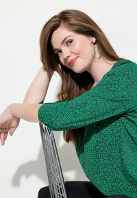 Zielona bluzka w kwiatowy wzór, z rękawami o długości trzech czwartych, zestawiona z akcentem srebrnego krzesła. Gładkie włosy spływające na ramiona.