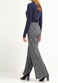 Mörkblå långärmad topp kombinerad med utsvängda, texturerade grå mönstrade byxor och mörkblå högklackade skor. Sedd bakifrån.