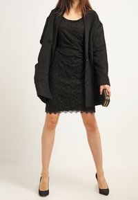 Vestido negro de encaje con una silueta ajustada, combinado con un abrigo negro suelto. Presenta un borde de encaje en el dobladillo y se acompaña de tacones altos negros.