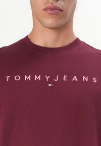 Burgunder T-Shirt mit Rundhalsausschnitt, mit "TOMMYJEANS" in pinker Stickerei und einem kleinen mehrfarbigen Logo. Weiche Stofftextur.