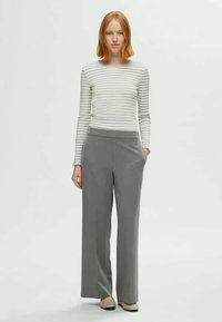 Jeune femme aux cheveux roux portant un haut rayé à manches longues, un pantalon large gris et des chaussures de couleur claire, se tenant avec une main dans la poche.