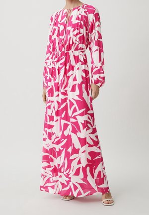 Femme portant une longue robe rose et blanche à motif floral, manches longues et taille froncée, associée à des talons blancs ouverts à l'avant.