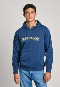 Modra kapuca iz mehkega materiala, ki ima sprednji žep, kapuco s vrvico in rumeno besedilo logotipa. Ujemajoče se z svetlimi jeans hlačami.