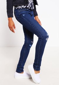 Osoba v tmavě modrých roztrhaných skinny džínách, bílých espadrilách a černé bundě s potiskem pod ní.