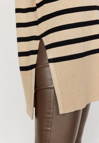 Beige gebreide trui met zwarte strepen, voorzien van een zijspleet, gecombineerd met glanzende bruine leggings. Getextureerde stof en aansluitend ontwerp.