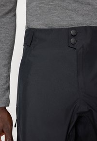 Close-up van een persoon die zwarte broek draagt met twee zwarte knopen en een grijs shirt met lange mouwen, waarbij een deel van de linkerarm en hand zichtbaar is.