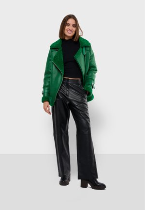 Giacca di pelle verde con collo peloso, abbinata a pantaloni larghi in pelle nera e stivali neri chunky con tacco. La modella è in posizione eretta.
