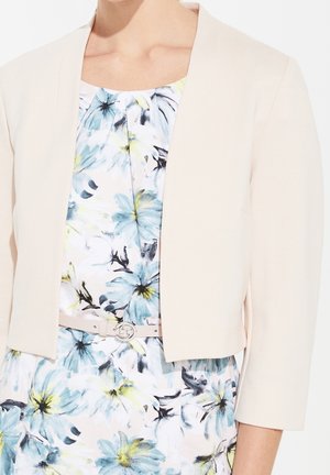 Femme portant un blazer court crème sur une robe blanche avec des motifs floraux bleus, noirs et jaunes, et une fine ceinture beige.