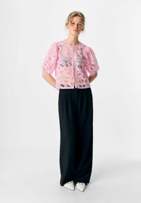 Une femme se tient avec les mains derrière le dos, portant un blouse en dentelle rose transparente, un large pantalon noir et des chaussures blanches à talons pointus, sur un fond uni.