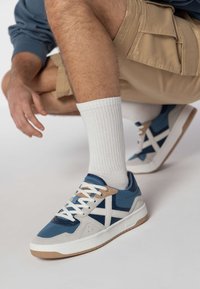 Modré a biele tenisky sú vyrobené z kombinácie sieťoviny a semišu, s gumovou podrážkou a hnedými akcentmi. Charakteristický krížový dizajn na boku.