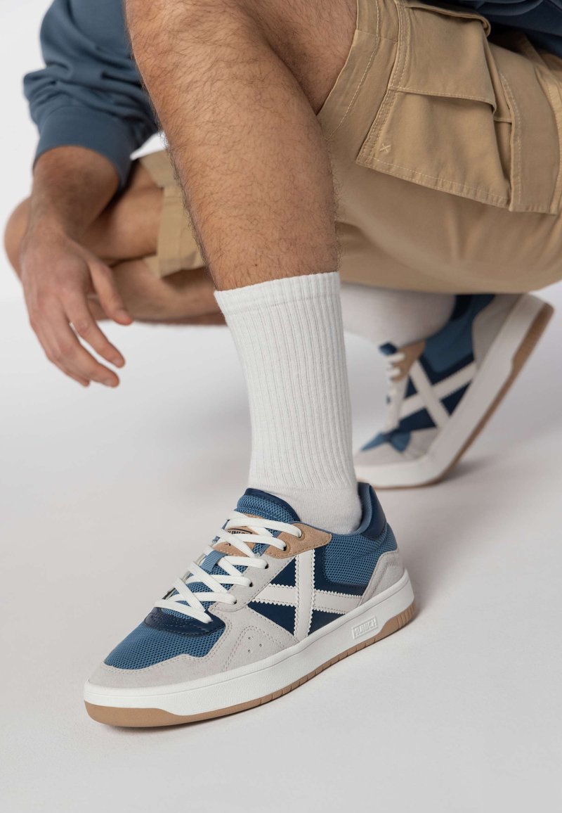 Modré a biele tenisky sú vyrobené z kombinácie sieťoviny a semišu, s gumovou podrážkou a hnedými akcentmi. Charakteristický krížový dizajn na boku.