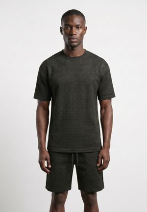Homme debout portant une chemise noire texturée à manches courtes et un short assorti sur un fond uni clair.
