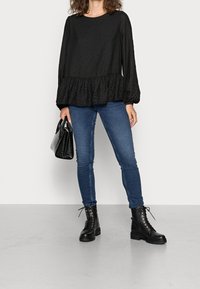 Femme portant une blouse noire transparente à manches longues avec un ourlet froncé, un jean skinny bleu, des bottes noires à lacets, tenant un sac à main noir sur un fond uni.