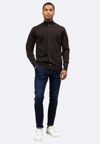 Cardigan marrone scuro con colletto alto, chiusura anteriore a bottoni e due tasche frontali, abbinato a jeans scuri e sneakers bianche.