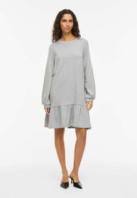 Robe grise en tissu doux, avec manches longues, encolure ronde et ourlet à volants. Le mannequin porte des talons noirs.