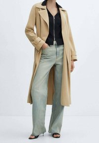 Beżowy trencz z dużymi klapami na czarnym topie na zamek, zestawiony z jasnoniebieskimi szerokimi dżinsami i czarnymi sandałami na obcasie.