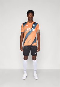 Narancssárga és fekete csíkos focimez „Leté” logóval, fekete rövidnadrággal, fehér zoknival és sportcipővel párosítva, atlétikai teljesítményre tervezve.