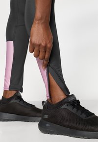 Svart atletisk leggings med pinke sideløsninger og lynlåse ved anklerne, parret med sorte mesh sneakers med en struktureret overflade og logo.