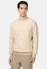 Pull à col roulé beige à motifs avec un design en diamants, fabriqué en matériau tricoté doux. Il présente des poignets et une taille côtelés, associé à un pantalon clair.