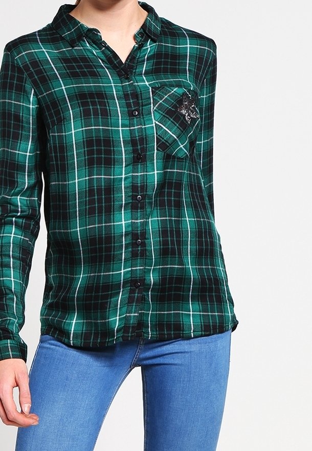 Femme portant une chemise à carreaux verte et noire à boutons avec une poche poitrine et un jean bleu clair sur un fond uni.