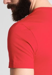 Camiseta de algodón roja con un diseño ajustado, mangas cortas y una textura suave. La costura del hombro es visible contra un fondo liso.