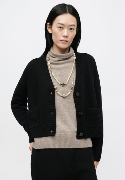 Μαύρη πλεκτή ζακέτα με ριμπ τελειώματα, διαθέτει κουμπιά και τσέπες. Φορεμένη πάνω από ένα ανοιχτό καφέ ζιβάγκο. Μακρυμάνικη, με άνετη εφαρμογή.