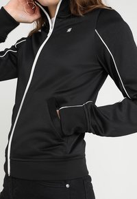 Chaqueta negra con cremallera, detalles en blanco, cuello alto, bolsillos laterales y tela texturizada. Diseñada para comodidad y uso activo.