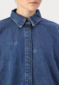 Denim jaka vidējā zilā krāsā ar pogu aizdari priekšpusē, norādītu apkakli un sirds formas izgriezumiem. Bojāta tekstūra un sudraba aksesuāri.