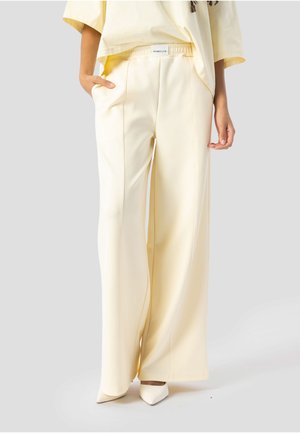 Kvinde iført cremefarvede bukser med vide ben og matchende cremefarvet top, den ene hånd i lommen, parret med hvide spidse sko, stående på grå baggrund.
