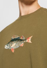 Muž nosící olivově zelenou košili s detailním vyšitým motivem ryby na horní části hrudi, detail výstřihu a ramene.