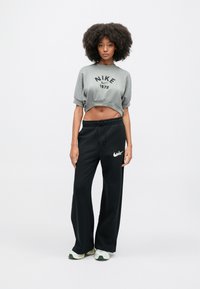 Siv pulover s kapuco, prirezan, z črnim napisom "Nike 1972", združen s črnimi širokimi hlačami s smrčkom Nike v beli barvi, nošen s supergami z zelenimi poudarki.