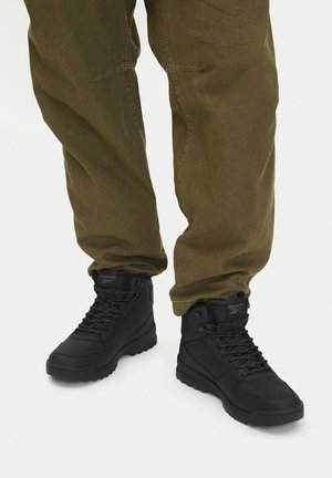 Czarne wysokie buty za kostkę z fakturowanej skóry, sznurowane, z gumowymi podeszwami, zestawione z oliwkowozielonymi, luźnymi spodniami.