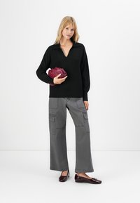 Mulher vestindo um suéter preto em V, calças cargo cinzas, sapatilhas burgundy, segurando uma mala de mão burgundy com franzido, em pé contra um fundo branco.