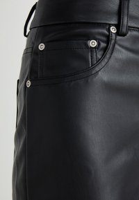 Jupe en cuir noir avec des accents en métal argenté, des coutures ton sur ton et une texture lisse, avec une vue rapprochée des poches et des détails.