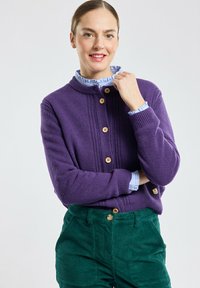 Cardigan viola lavorato a maglia con scollatura rotonda, bottoni in legno e texture a coste, abbinato a pantaloni in velluto a coste verdi e una camicia blu con colletto.