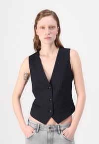 Gilet a righe nere con scollatura a V profonda, cinque bottoni e tasche laterali, abbinato a jeans grigi chiari. Texture del tessuto liscia.