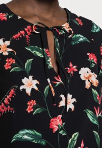 Černá květinová blůza s výstřihem ve tvaru klíčové dírky a mašlí. Má živé květiny v červené, oranžové a bílé barvě s zelenými listy na měkké tkanině.