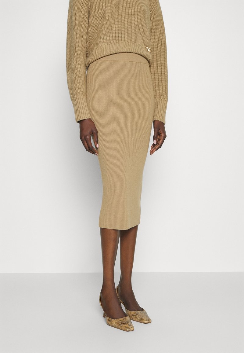 Femme portant une jupe midi côtelée beige en maille et un pull assorti, avec des escarpins pointus à imprimé serpent, debout devant un fond blanc.