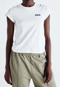 Bílá bavlněná tričko s krátkými rukávy a srolovanými okraji, na hrudi má černou nášivku s logem Levi's, v kombinaci se světle olivově zelenými joggery.