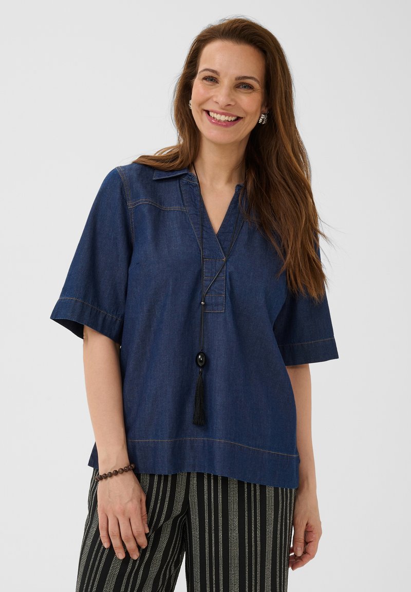 Femme souriante aux longs cheveux bruns portant un haut bleu foncé à manches courtes, un pantalon rayé noir et blanc, un collier à pompon noir et un bracelet de perles.