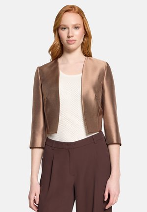 Young woman with red hair wearing a bronze cropped satin blazer over a white textured top and brown high-waisted trousers.