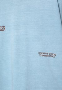 Helles blaues T-Shirt mit kurzen Ärmeln aus weichem Stoff. Verfügt über braun bedruckte Texte an zwei Stellen: "CREATIVE STUDIO" und "AUSSTELLUNG."