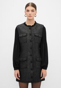 Vestido de tweed negro con escote fruncido, cuatro bolsillos y botones en la parte delantera. Mangas largas negras con textura plisada. Tejido suave y texturizado.