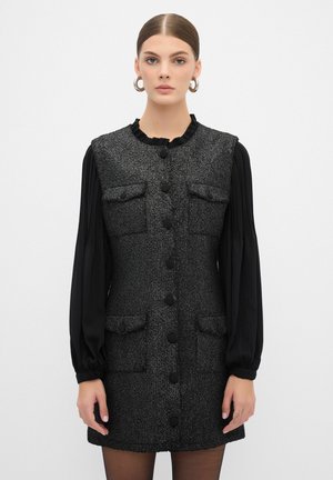 Musta tweed-mekko, jossa on röyhelökaulus, neljä taskua ja edessä napit. Pitkät mustat hihat, joissa on pleat-tekstuuria. Sileä, teksturoitu kangas.